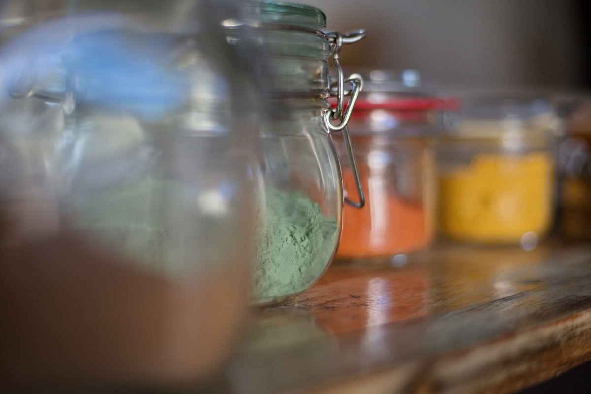 Makró részletfotó egy fa felületen sorba rendezett üvegekről, amelyekben sárga, terrakotta és mentazöld tónusú természetes földpigment porok láthatók a műteremben.