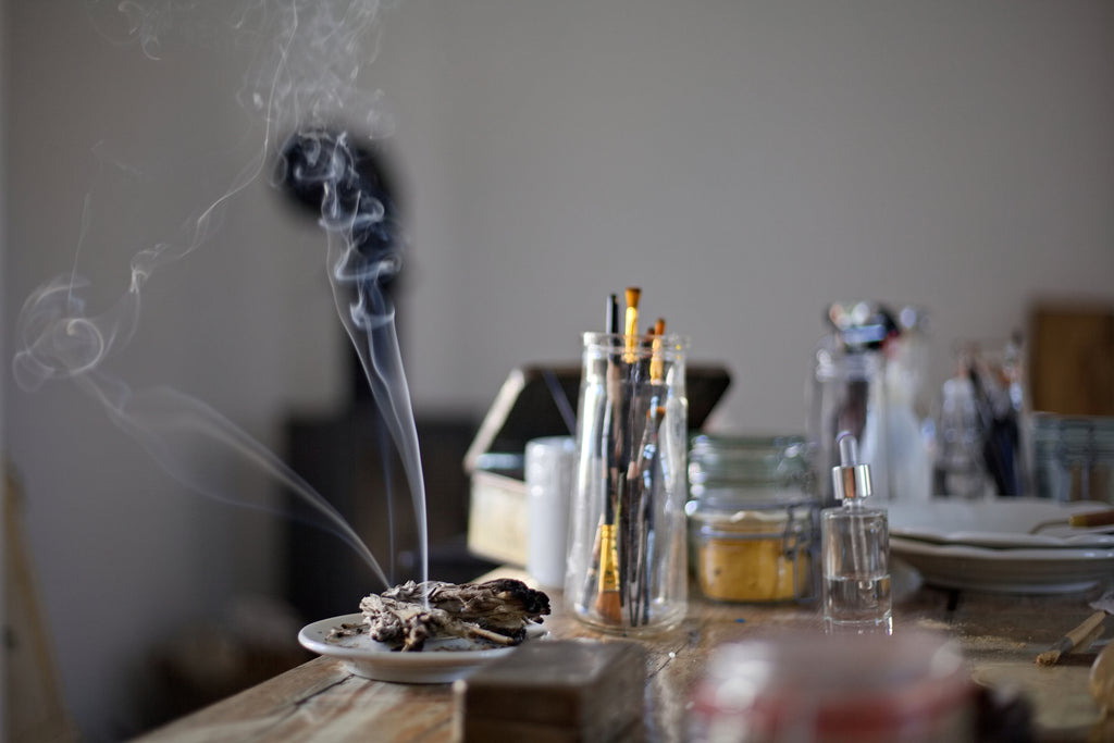 Közeli kép egy műtermi rituáléról, ahol egy sárgaréz tartóból gomolygó füstölő füstje száll fel a fa asztalon lévő ecsetek és földpigmentes üvegek között.