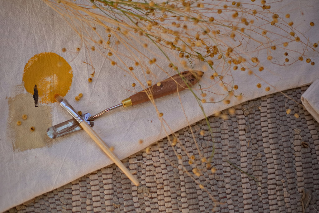 Makro fotó szőnyegre terített khadi anyag részlet, rajta a spatula és a kézzel festett földpigment minta és egy száraz virág